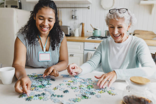 portrait d’une travailleuse sociale bénévole afro-américaine jouant à un jeu de puzzle avec une dame âgée joyeuse, s’amusant, riant à la recherche de la bonne pièce assise ensemble à la table de la cuisine - jeu personne agée photos et images de collection